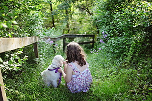 girl wearing white and purple dress playing with white small dog in garden beside fence during daytime