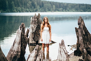 selective focus photography of woman in white floral mini dress standing on body of water surrounded by tree trunks