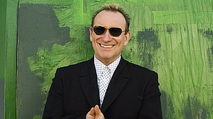 men's black suit and gold framed sunglasses standing in front of green painted wall during daytime