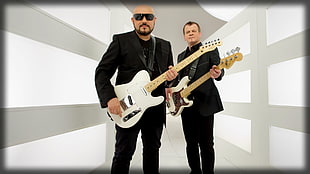 photo of man wearing black sunglasses, suit jacket, and pants playing white electric guitar near man playing guitar