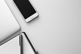 white android smartphone on top of white surface near the notebook with pen on top