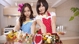 two women standing wearing blue and red aprons holding tray