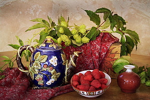 red strawberry on red and white ceramic bowl beside blue, green, and white floral ceramic teapot and green petaled flower
