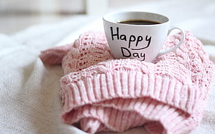 white ceramic Happy Day printed teacup filled with coffee on pink knit shirt