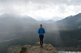 woman in blue pullover hoodie standing on the stone cliff on the mountain during daytime