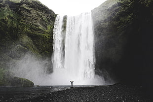 person standing facing waterfalls under white sky