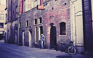 man in blue shirt and blue denim jeans leaning on concrete brick wall building