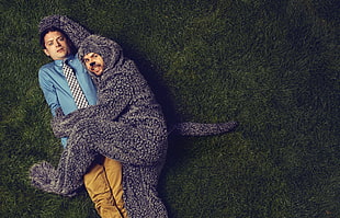 man with mascot hugging man wearing blue shirt while laying on green grass field