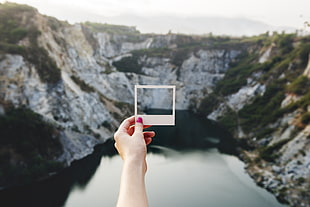 woman holding photo with mountain view near river at daytime