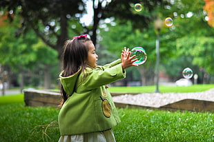girl in green hoodie standing on green grass field trying to catch bubble