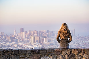 photo of blonde haired girl in brown jacket sitting on concrete stone platform facing in front of high rise buildings