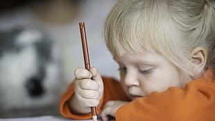closeup photo of girl in orange long-sleeved shirt holds pencil