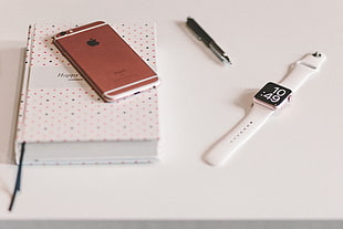 rose gold iPhone 6s on white book and rose gold aluminum case Apple Watch with white Sports Band beside mechanical pen