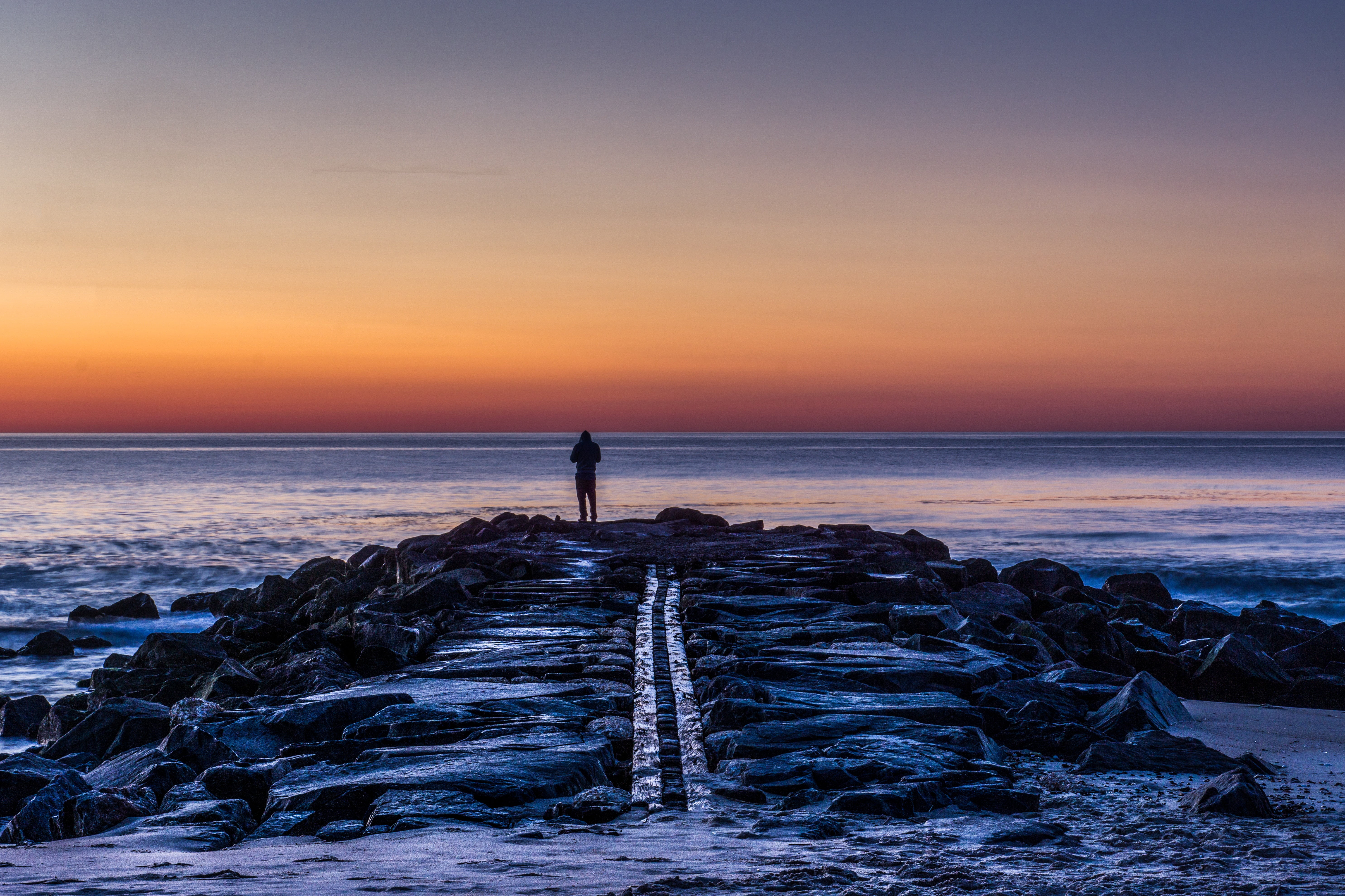 silhouette photo of person standing on rock facing sea during sunset