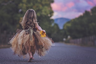 girl holding yellow sunflower walking away from the camera on asphalt road and forest tree photography