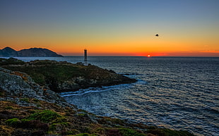 white and black lighthouse near body water and airplane flying over the sunset
