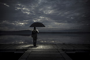 grayscale photography of man in white top holding umbrella near body of water