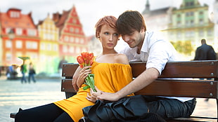 selective focus photography of man sitting on brown bench in front of woman holding orange petaled flower