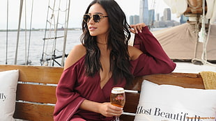 woman wearing red deep-plunging cold shoulder long-sleeved dress holding glass of cocktail while sitting on wooden bench inside boat