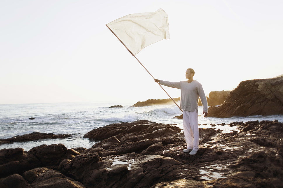 man wearing gray long-sleeve shirt holding white flag standing near ocean during daytime HD wallpaper