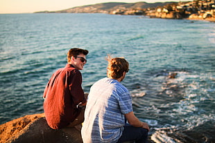 two men sitting on the rock near on the sea during daytime HD wallpaper