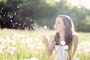 woman in white sleeveless top blowing dandelion during daytime