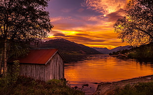 brown wooden shed near body of water and mountain at sunset