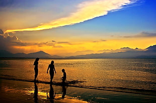 silhouette of three person on seashore under white clouds and blue skies at sunset HD wallpaper