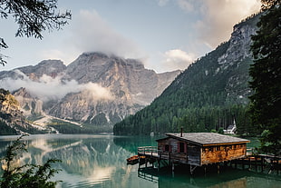 landscape photography of brown wooden house on top of body of water facing tall mountains surrounded of tall tres