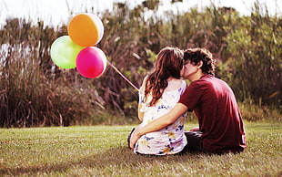 couple kissing while holding balloons and sitting on grass during daytime