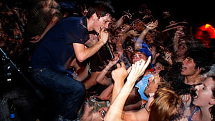man in black shirt and blue denim jeans holding microphone singing in front of crowding people
