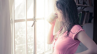 woman wearing pink shirt drinking coffee looking outside window