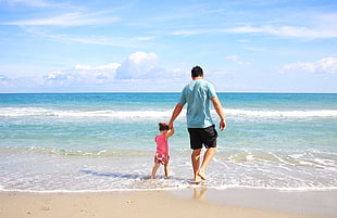 man in blue t-shirt holding hands with girl in pink tank top while walking on beach shore during daytime