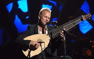 shallow focus photography of man in black notch lapel suit jacket playing brown and black string instrument in front of black microphone with stand