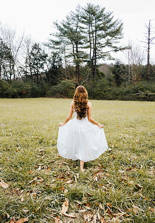 girl in white dress standing on grass field while facing on tall trees at day time