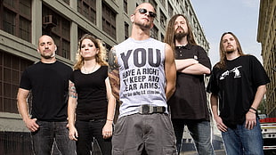 man in white sleeveless shirt with arm tattoo wearing gray pants and four people behind wearing black shirts near high-rise building digital wallpaper