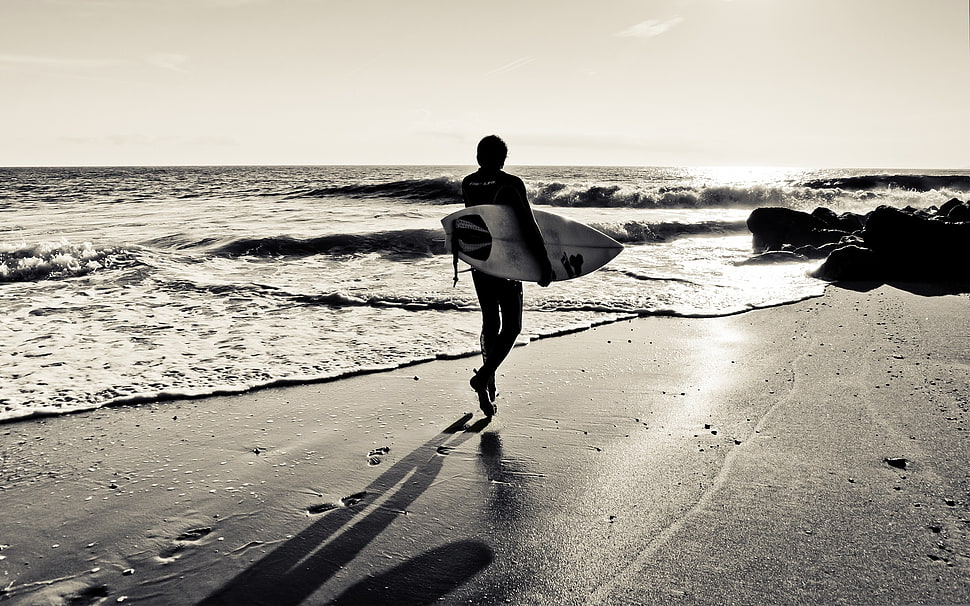 black and white photograph of person holding surfing board while walking on sea shore HD wallpaper