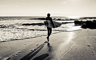 black and white photograph of person holding surfing board while walking on sea shore