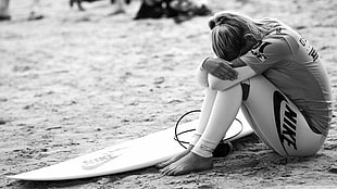 grayscale photography of woman sitting on sand her beside surfboard