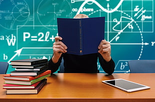 man in black longsleeve shirt holding blue book sitting on brown wooden desk
