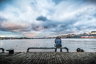 person in gray jacket and blue backpack sitting on black steel tube bench beside body of water under white clouds HD wallpaper