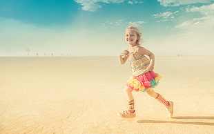 panoramic photo of girl wearing striped mini dress running on beige sand field under white clouds while smiling, kid