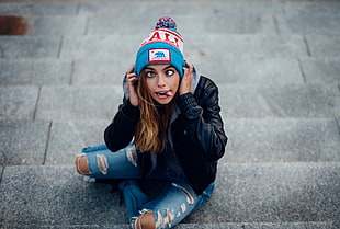 woman wears black leather jacket and distressed blue denim jeans sitting on concrete stairs making faces while holding on bobble hat