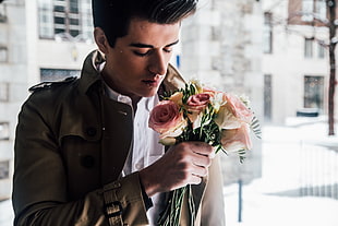 man in brown jacket holding while smelling pink flowers