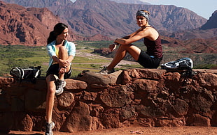 two woman sitting on rock formation wall during daytime