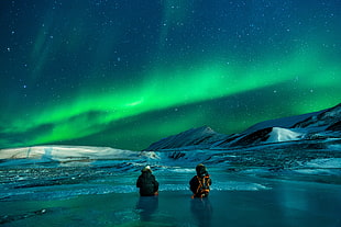 two people sitting on ice field under aurora borealis