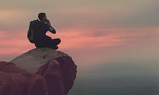 man wearing black cloth and hiking backpack sitting on rock while meditating during daytime