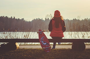 woman sitting on bench beside red long board in front of lake