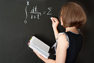 brunette short-haired woman wearing black sleeveless top holding notebook writing math equation on blackboard