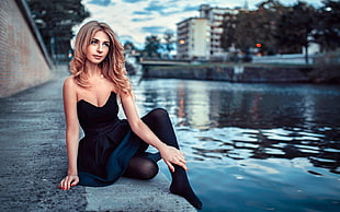 woman wearing black strapless dress sitting beside body of water at daytime